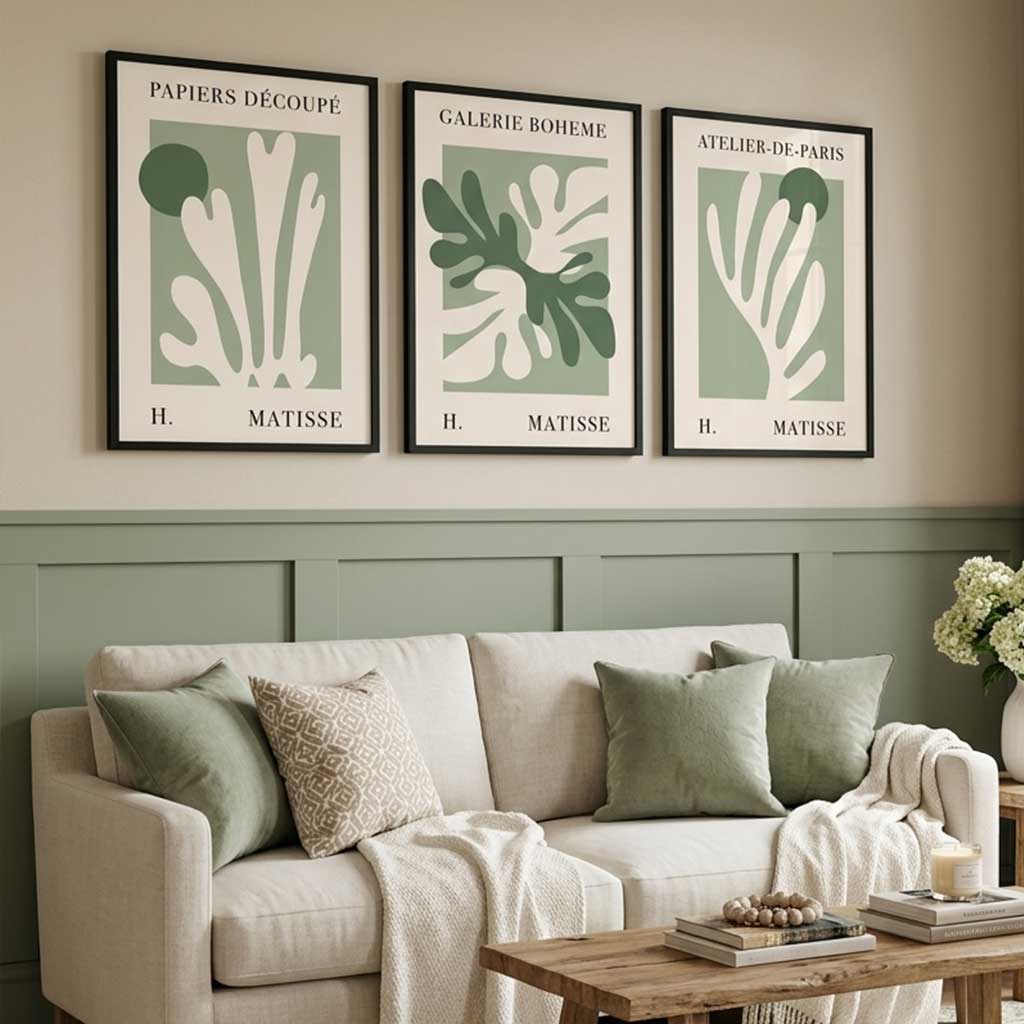 Three framed wall art prints with minimalist botanical designs in sage green and cream hang above a beige sofa in a styled living room with green wainscoting, pillows, a wooden coffee table, and a vase of flowers.