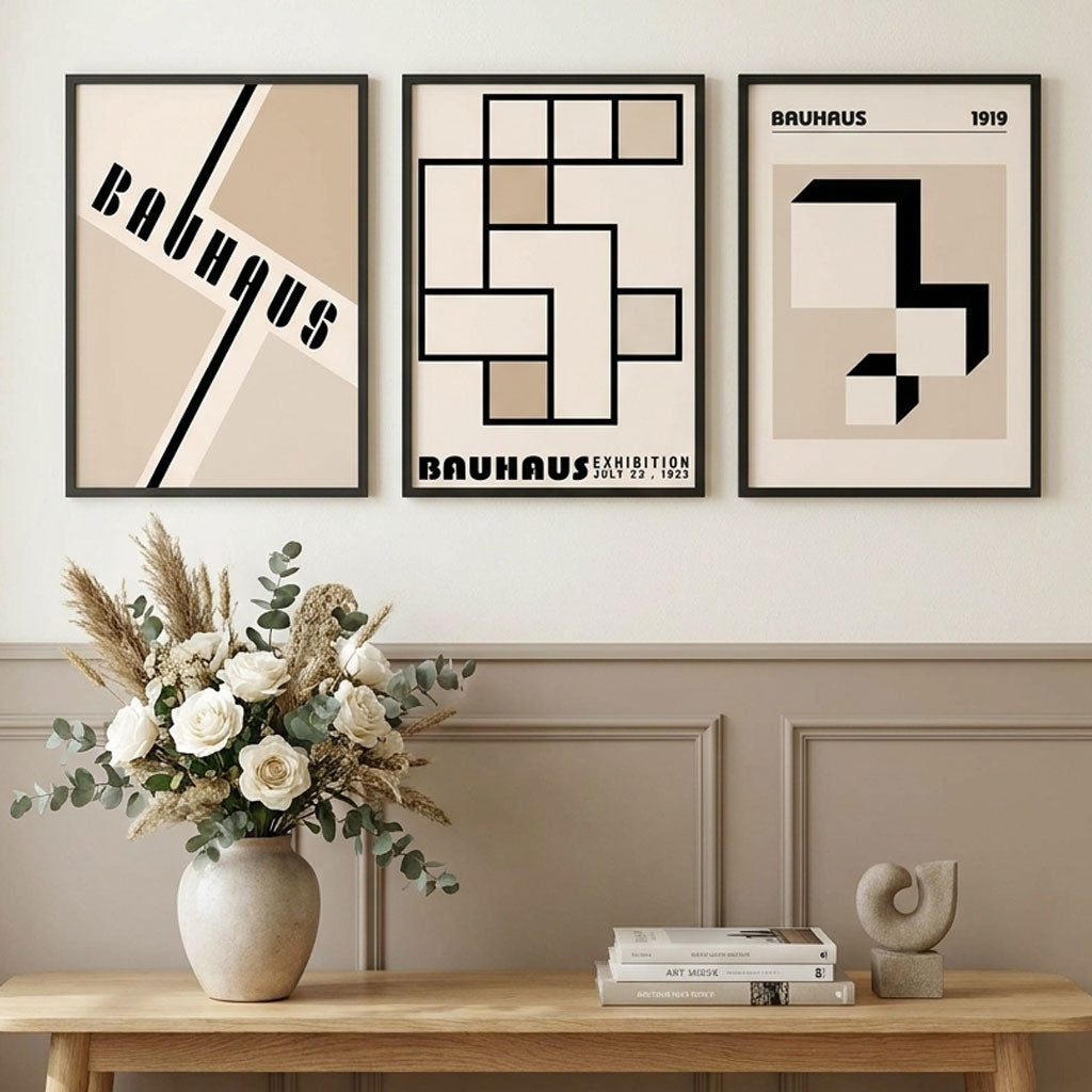 Three Bauhaus-themed posters on a wall with a vase of flowers and books on a table below.