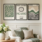 Three framed wall art prints hang above a cream armchair in a bright living room. The prints feature abstract and vintage-inspired designs in black, white, and muted green, with text including 'MUSEE D'ANTIBES', 'PICASSO', and 'BAUHAUS'.