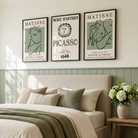 A styled bedroom scene with a bed dressed in cream bedding and a textured olive green throw blanket, set against a light green paneled wall. Above the bed are three framed posters featuring Matisse and Picasso.