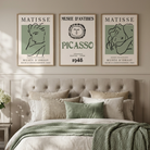 A styled bedroom scene with a bed dressed in neutral linens and a green knit throw, beneath three framed wall art prints featuring Matisse and Picasso. A small side table with white flowers sits beside a window, and the room has beige walls, wood flooring, and a tufted headboard.