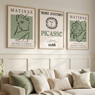 Three framed vintage-style wall art prints with Matisse and Picasso text hang above a light beige sofa in a bright living room with cream walls, wood wainscoting, a side table with flowers, and a window with sheer curtains.