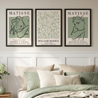 Three framed Matisse-style wall art prints hang above a bed in a softly styled bedroom. The prints feature muted sage green and cream abstract line-art designs with black-framed borders.