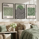 Three framed Matisse, Kusama, Bauhaus wall art prints with green, black, and cream abstract and typographic designs hang above a bed in a softly lit bedroom. The room includes beige bedding, pillows, a bench with green and beige cushions, and a vase of white flowers.