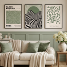 Three framed abstract art prints in black, cream, and sage green hang on a beige wall above a cream sofa with green pillows and a beige throw blanket in a styled living room.