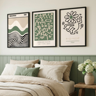 Three framed abstract art prints hang above a bed in a styled bedroom. The prints feature minimalist black, green, beige, and white designs, including wavy lines, geometric patterns, and a flower motif.