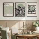 Three framed art prints with minimalist Japanese and abstract designs are displayed on a beige wall above a round wooden dining table with green and beige chairs in a styled dining room.
