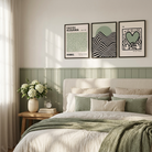 A styled bedroom scene with a bed dressed in white and sage green bedding, a wooden side table with a vase of white flowers, and three framed wall art prints above the headboard. The room has a light neutral wall, window-side curtains, and a sage green wainscoted panel.