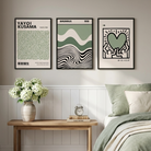 Three framed wall art prints with black, white, and sage green designs hang above a wooden bench in a softly styled bedroom with green bedding, pillows, a vase of flowers, and a window.