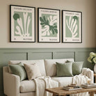 Three framed wall art prints with minimalist botanical designs in sage green and cream hang above a beige sofa in a styled living room with green wainscoting, pillows, a wooden coffee table, and a vase of flowers.